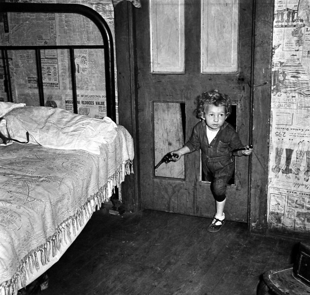 Bonjour Papa Bonjour Maman. Coal miner’s child using a hole in the door to enter a bedroom with a smoking pipe in one hand and a gun in the other in Bertha Hill, West Virginia. Photo by Marion Post Wolcott. 1938