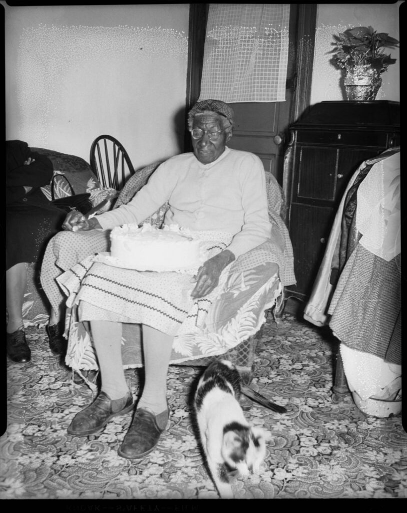 les esclaves se reposent le dimanche. Charles « Teenie » Harris Portrait of former slave Sabre “Mother” Washington, holding cake on her lap, on her 109th birthday, in her home on Conemaugh Street with cat in foreground, 1954