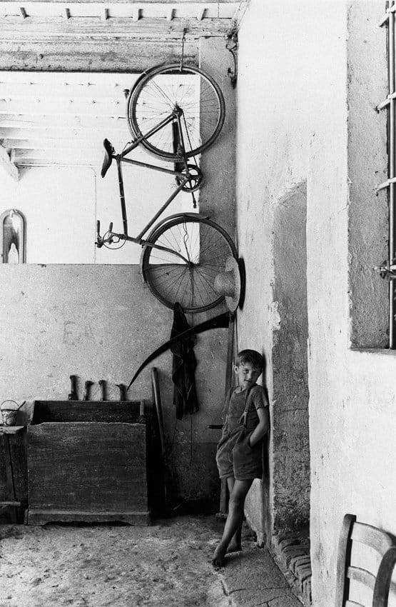 un jeune garçon désoeuvré appuyé contre un mur, les mains dans les poches, à côté de son vélo suspendu au plafond de l'atelier