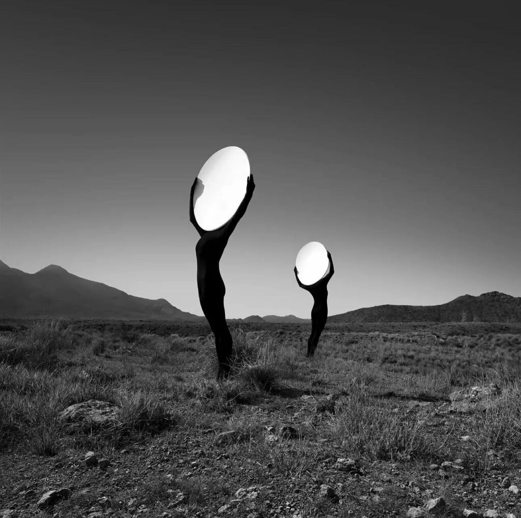 dans un paysage aride, deux femmes en justaucorps noir tendant vers le ciel devant leur visage un grand miroir ovale
