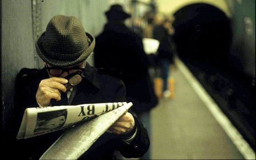 sur le quai du métro, un homme en pardessus et chapeau mou, portant déjà des lunettes, scrute avec des verres grossissants la une du journal