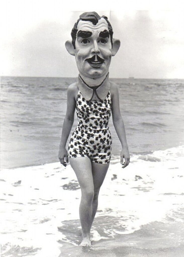 une femme en maillot de bain, portant un masque d'homme à moustache, sort de l'océan