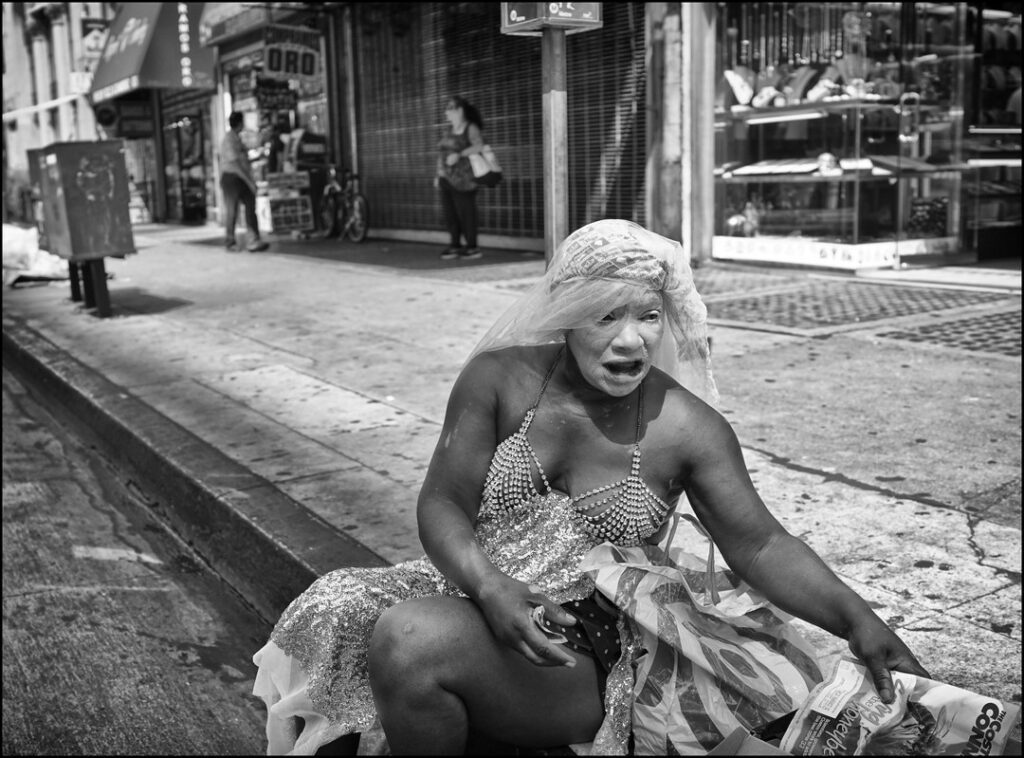 un homme noir en robe du soir, le visage et les cheveux blanchis, la bouche ouverte comme dans un cri, assis dans la rue sur le bord du trottoir