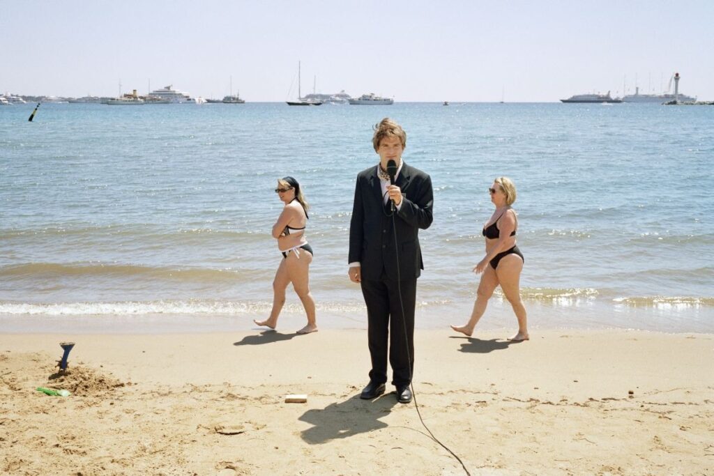 un journaliste en costume tenant son micro debout sur la plage ensoleillée, deux femmes passent en maillot de bain derrière lui, au loin flottent les yachts