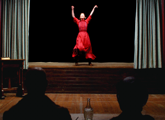 une femme en robe rouge, le sourire aux lèvres, exécute sur scène une chorégraphie sommaire de célébration de victoire à base de frappe des pieds sur le sol et de bras alternativement levés
