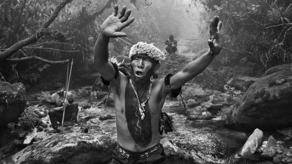 Au centre de cette photo en noir et blanc, on voit un homme, des cuisses jusqu'aux épaules, debout avec les bras levés, paumes tournées vers l'avant. Sa bouche est ouverte, comme s'il chantait ou psalmodiait. Il est torse nu, mais porte un chapeau fait de plumes, et ses joues et son front sont recouverts de peinture sombre. La peinture descend sur son torse, pour s'arrêter près de son nombril. Un collier pend sur sa poitrine.
Derrière l'homme, on peut voir un cours d'eau peu profond qui s'écoule sur des rochers, et des branches d'arbres luxuriantes qui s'arc-boutent au-dessus. Deux autres hommes sont présents à l'arrière-plan. L'un d'eux est assis sur un rocher recouvert de mousse à gauche de l'image, et tient deux longues lances. L'autre se trouve en haut au centre de l'image, accroupi sur une butte. Un épais brouillard flotte dans l'air.