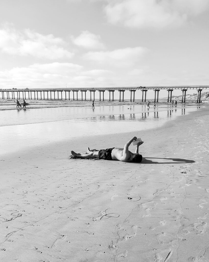 allongé sur le dos sur la plage, un homme tient devant le ciel son livre à bout de bras 