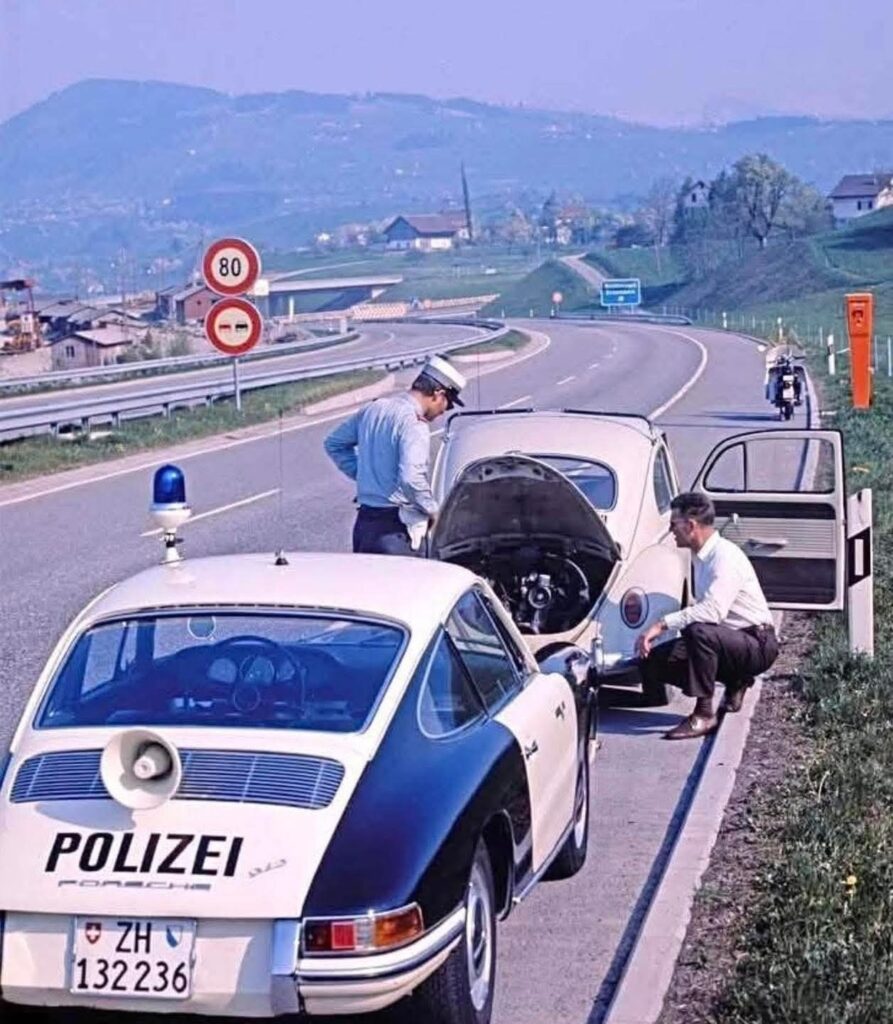 un agent de la police suisse (canton de Zurich) de l'autoroute se penche sur la détresse d'un usager agenouillé près de sa Volkswagen en panne garée sur la bande d'arrêt d'urgence, le capot levé