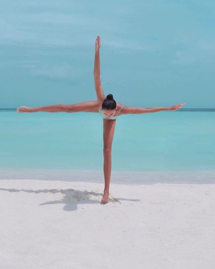 une femme en croix sur la plage, bras gauche et jambe droite parallèle à la ligne d'horizon de la mer, jambe gauche en appui sur le sable, bras droit tendu vers le ciel