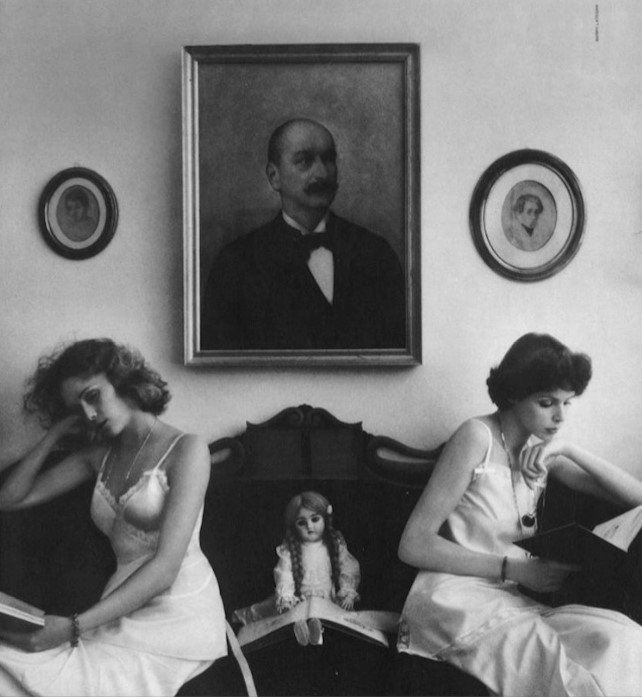 sur un canapé, deux jeunes femmes et une poupée, au dessus d'elles, le portrait du père moustachu dans son cadre en bois