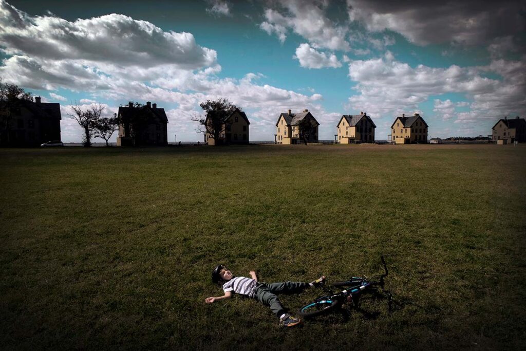 allongé sur le dos sur une vaste prairie devant une rangée de maison, un jeune garçon et son vélo face au ciel