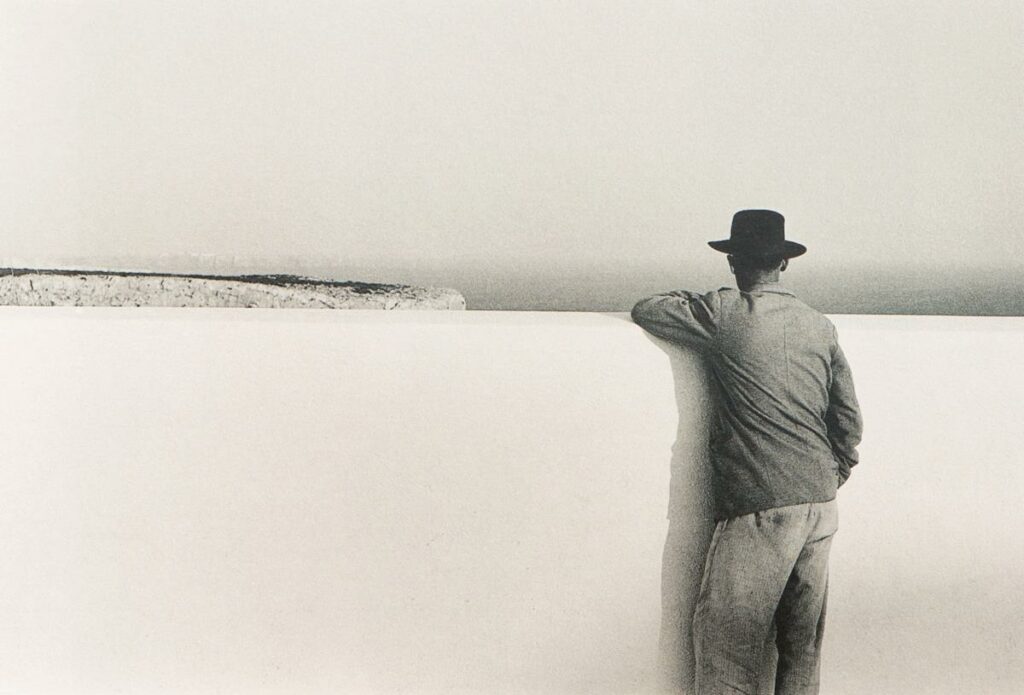 le bras appuyé sur un mur blanc lui arrivant aux épaules et partageant l'image horizontalement en deux, un homme de dos regarde ce qui est de l'autre côté, une falaise, la mer, l'horizon