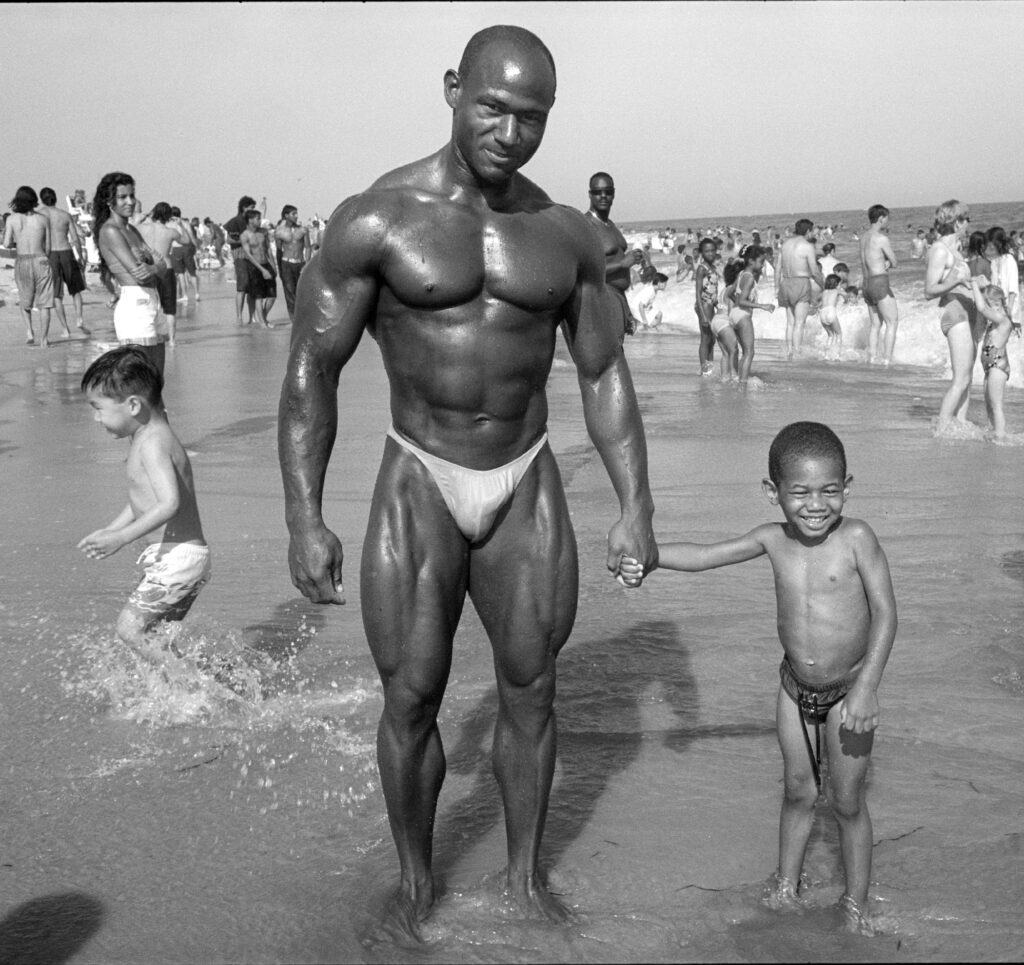 sur la plage, les pieds dans l'eau, un petit garçon souriant donne la main à son père culturiste aux muscles puissants luisant au soleil