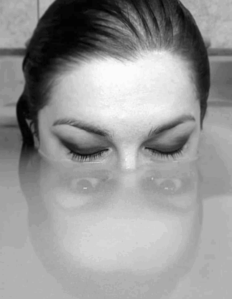 le bas du visage immergé, une femme aux yeux clos les a grand ouverts dans son reflet sous la surface