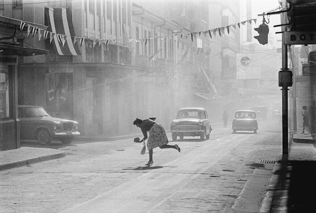 Une atmosphère brumeuse, chargée de cendres volcaniques, enveloppe la ville de San José. Une femme traverse la rue en titubant à cause de la densité du brouillard, tandis que les voitures se frayent un chemin à tâtons. Les cendres dévastatrices, qui recouvrent littéralement tout dans la ville, sont le résultat de l'éruption actuelle du volcan Irazu, habituellement inactif, à une trentaine de kilomètres de là.