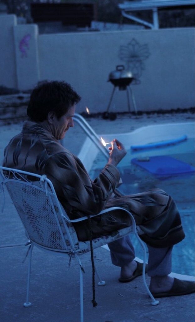 dans la pénombre, assis en robe de chambre dans un fauteuil de jardin devant la piscine, Walter White regarde la flamme d'une allumette