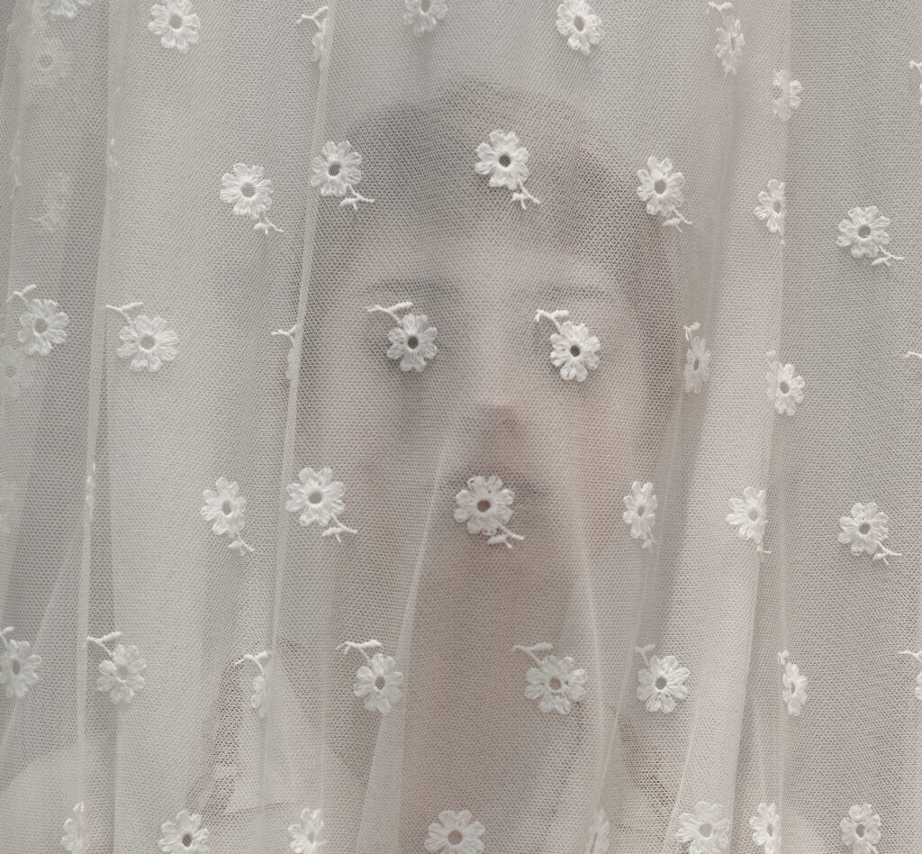 le visage d'une femme derrière un rideau, le motif floral du voilage coïncide avec sa bouche et ses yeux
