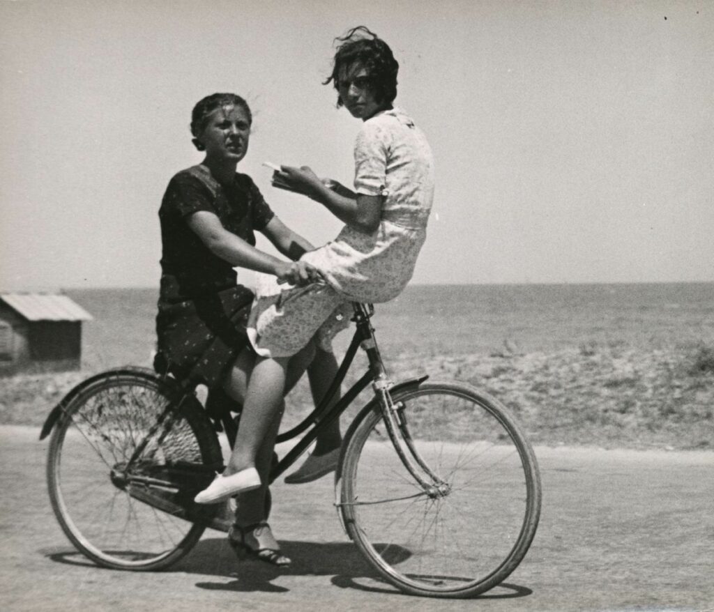 sur une route de campagne, deux femmes face à face sur une bicyclette, l'une assise sur le guidon
