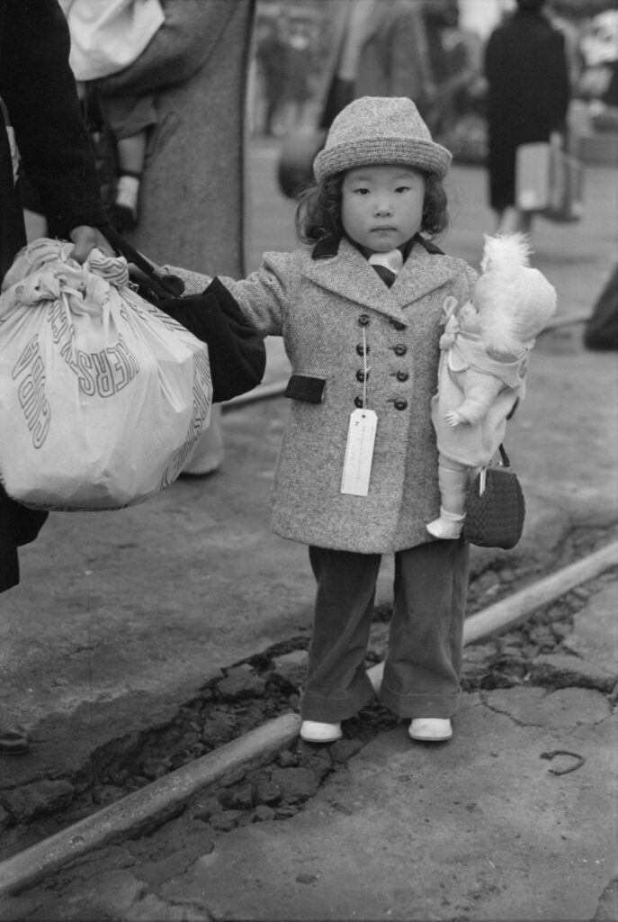 une petite fille nippo-américaine affublée d'une étiquette donne la main à sa mère chargée de bagages, attendant avec ses parents d'être déportée vers un camp de la vallée d'Owens, Los Angeles, 1942