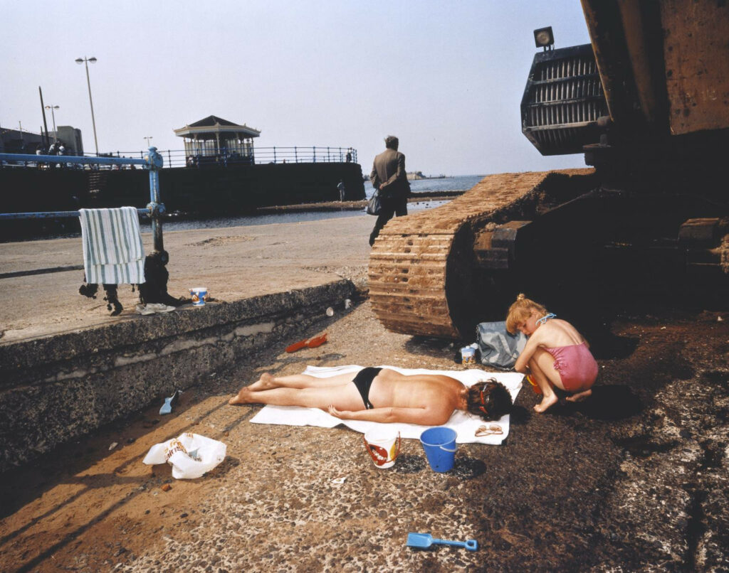 au bord de la mer, à quelques centimètres des chenilles d'un engin de chantier, une femme se fait bronzer sur le béton de la jetée