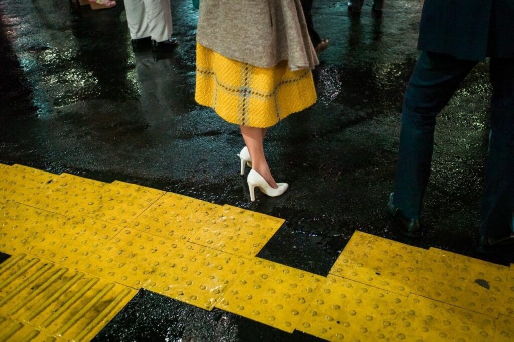 une femme portant une jupe à damier jaune attend sur le quai, un carré de même couleur manque à la bande podotactile pour malvoyants