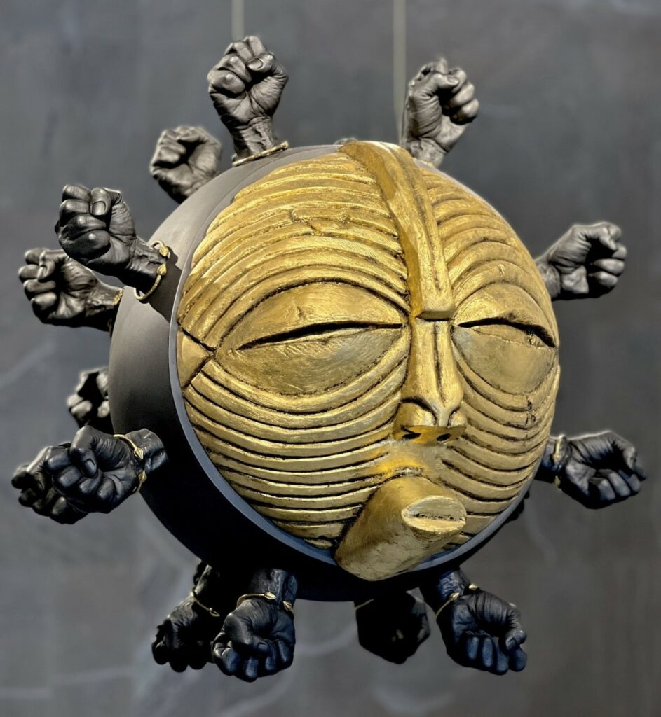 comme un masque africain ou un virus, la face d'or d'une sphère noire aux yeux plissés et aux lèvres tendues, hérissée d'une multitude de poings serrés