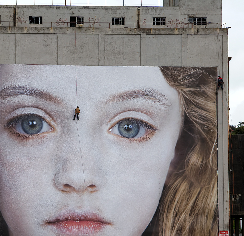 installation sur la façade d'un immeuble d'un panneau géant représentant le visage d'une jeune fille ; entre ses yeux, un homme suspendu en rappel