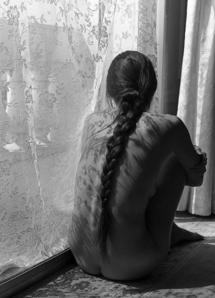 assise les bras noués autour de ses genoux, une femme nue aux cheveux tressés en une longue natte, attend derrière les rideaux blancs