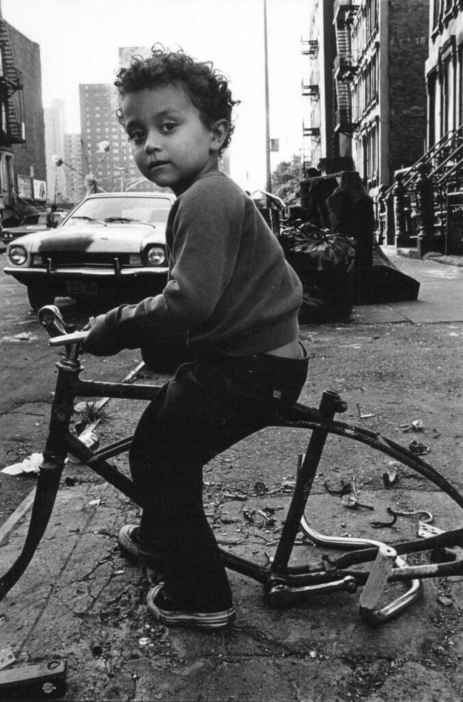 dans une rue de la ville, portrait d'un enfant juché sur le cadre d'une épave de vélo sans selle ni roues