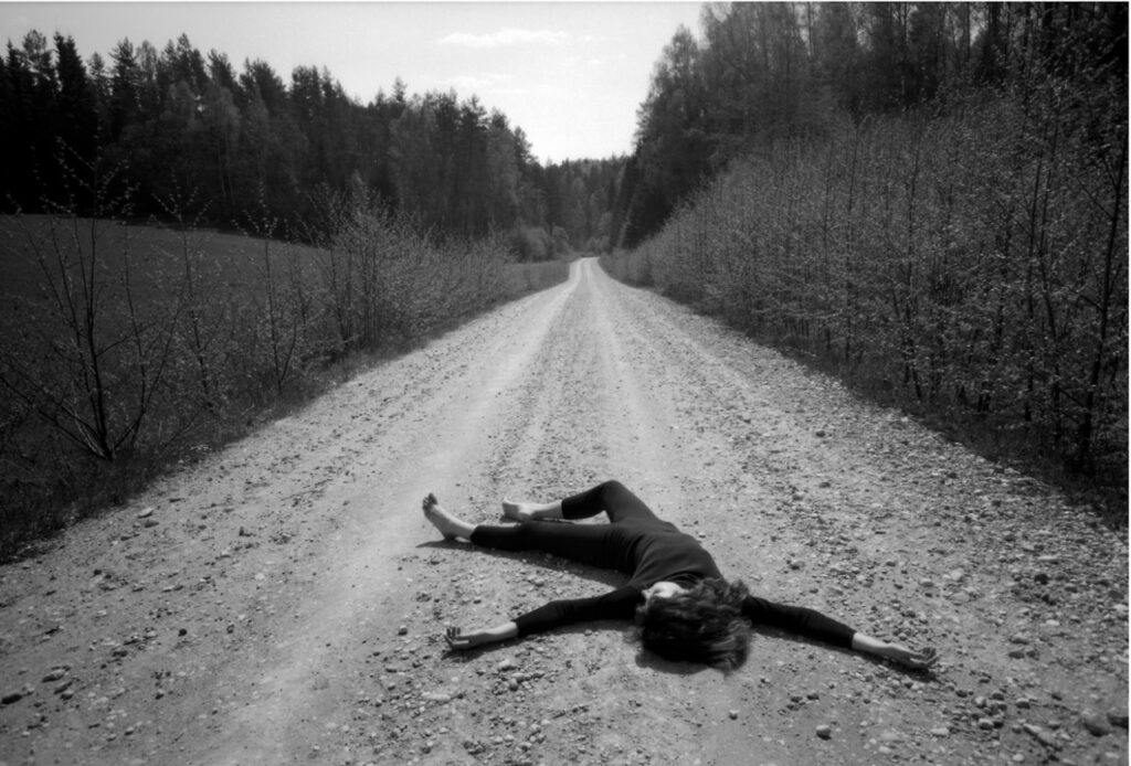 une femme en noir, pieds nus, allongée sur un chemin de terre bordé de haies traversant une forêt de résineux