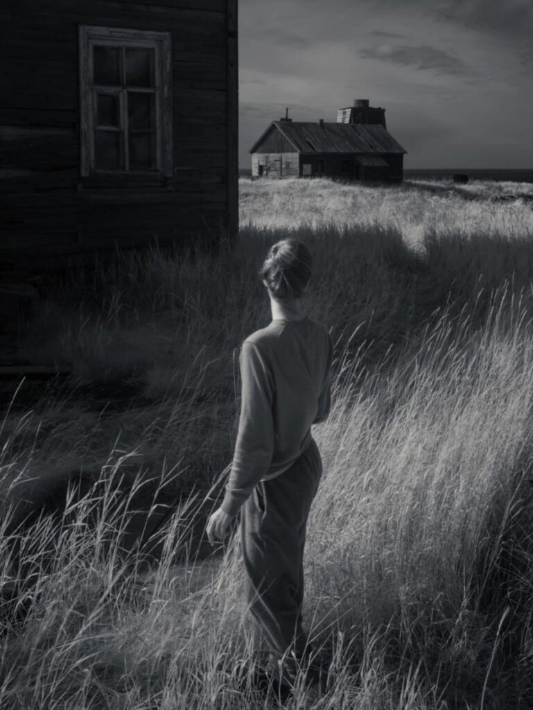 une femme dans les hautes herbes derrière la maison abandonnée, le regard tourné vers le large