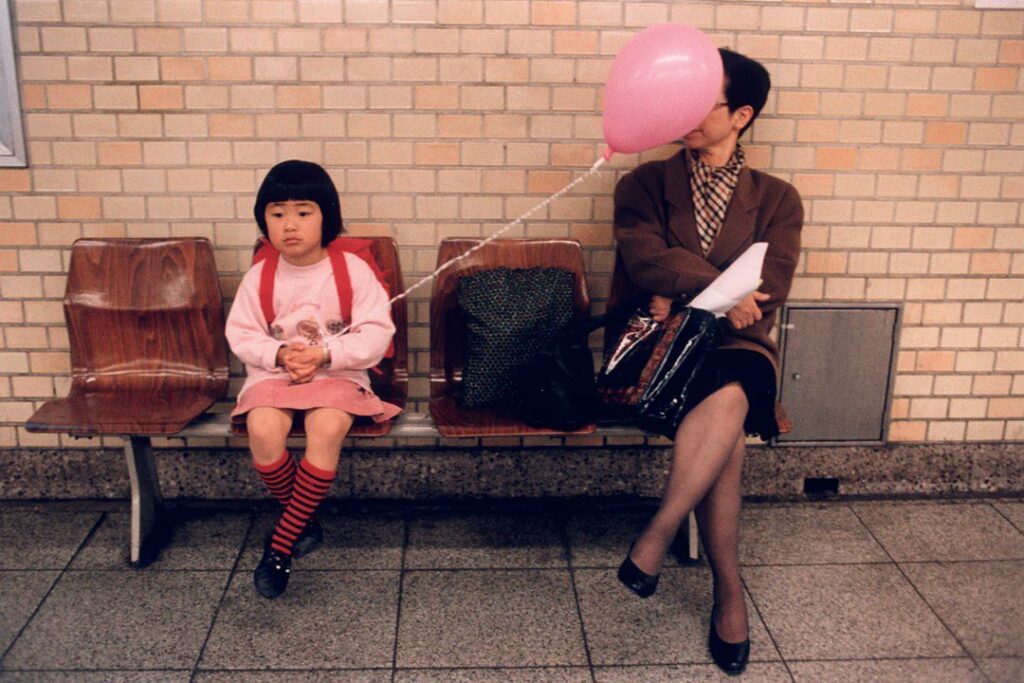 sur un banc public, une petite fille indifférente au fait que son ballon rose cache le visage d'une femme adulte assise à côté d'elle