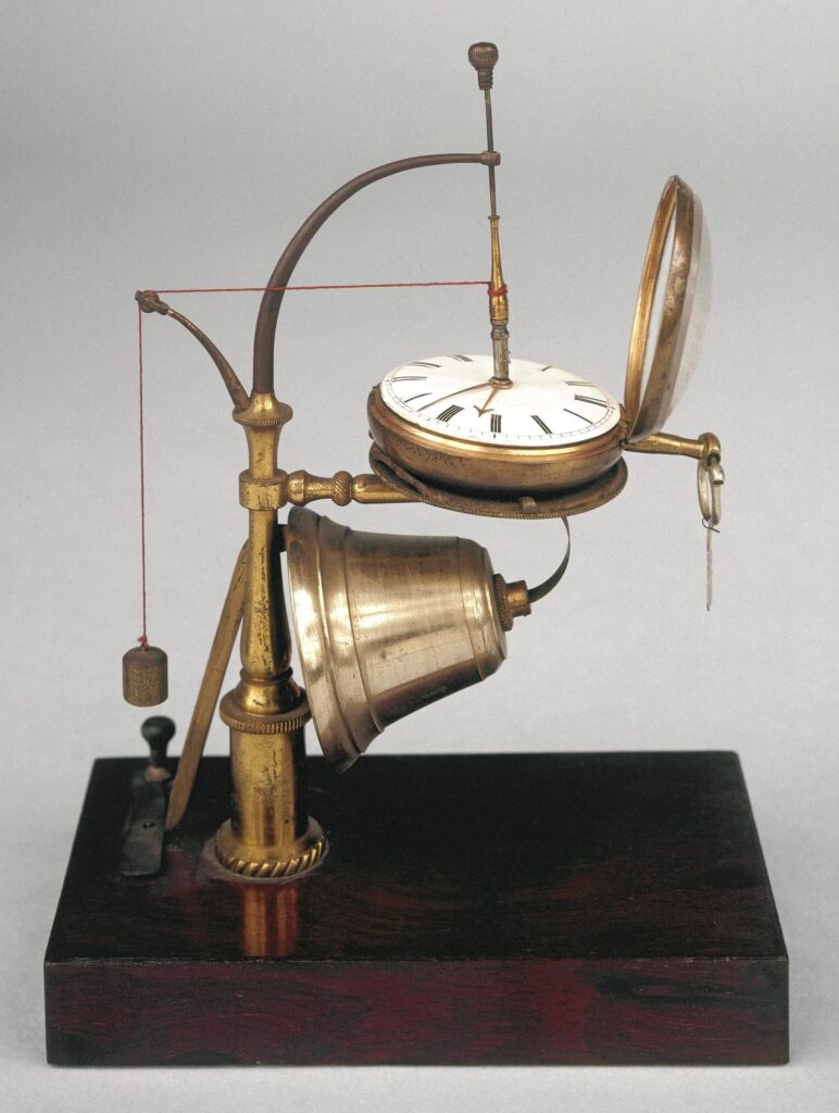 Une colonne verticale en laiton tourné, montée sur un socle rectangulaire en acajou, supporte un porte-montre horizontal sous lequel est fixée une cloche à ressort. La cloche est actionnée par un levier maintenu en place par un ressort fixé au socle. Un cordon lesté, partant de l'axe central de la montre, fonctionne comme un piège à souris : lorsque le lest touche le ressort, la cloche se déclenche