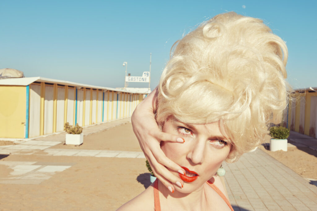 sur les planches au bord de la mer, entre deux rangées de cabines de bain, une femme blonde à la coiffure élaborée passe son bras gauche derrière sa tête pour poser son annulaire dubitatif sur la commissure droite de sa bouche rouge