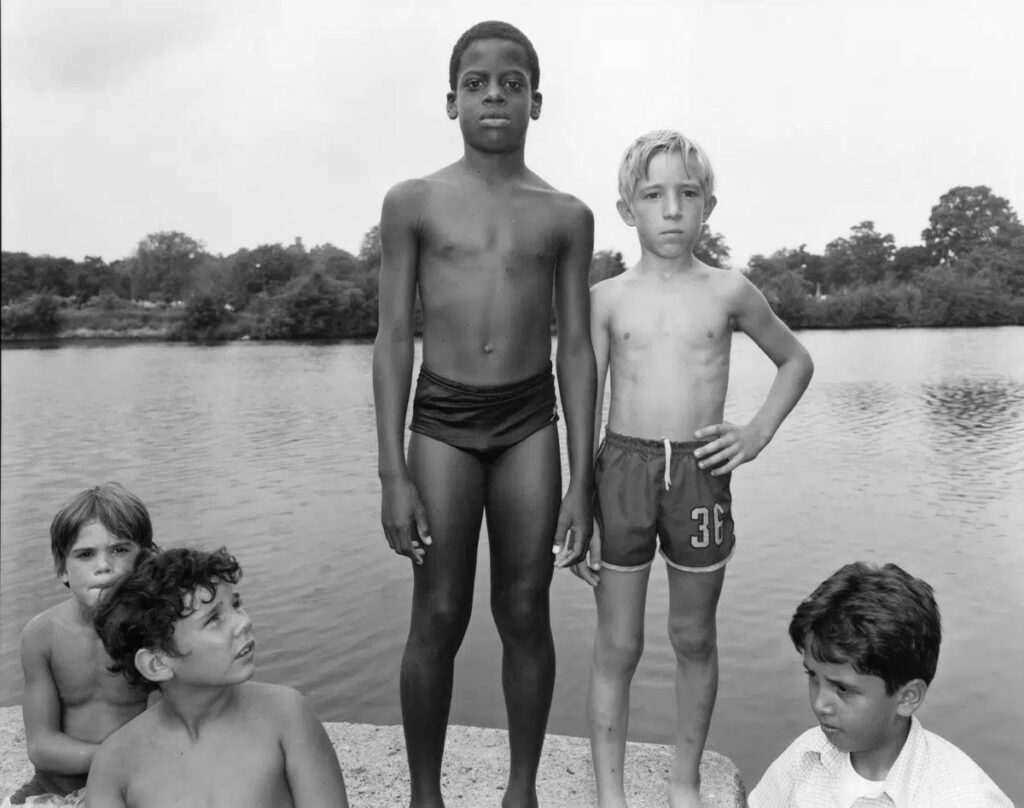 en maillot de bain sur un quai au bord d'une rivière, quatre garçons blancs ; l'un d'eux, debout, se tient à côté d'un garçon noir