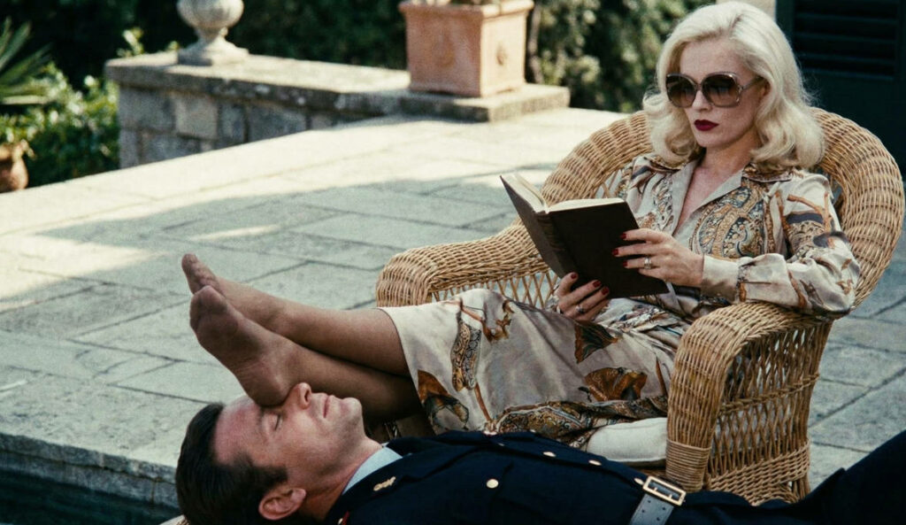 assise dans un fauteuil en rotin, Emmanuelle lit, les pieds croisés sur le visage d'Emmanuel en uniforme