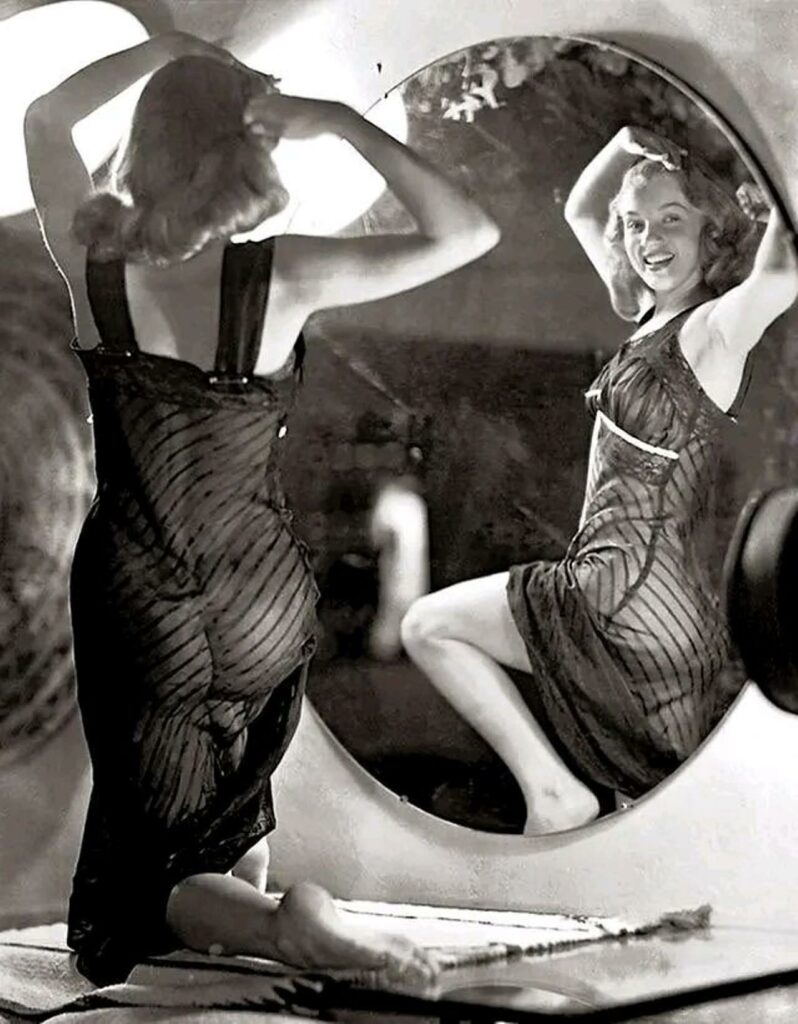 Marilyn Monroe portant un déshabillé transparent posant devant de trois-quarts dos devant un grand miroir, prétendant parfaire sa coiffure et offrant l'image de son corps de starlette aux voyeurs 