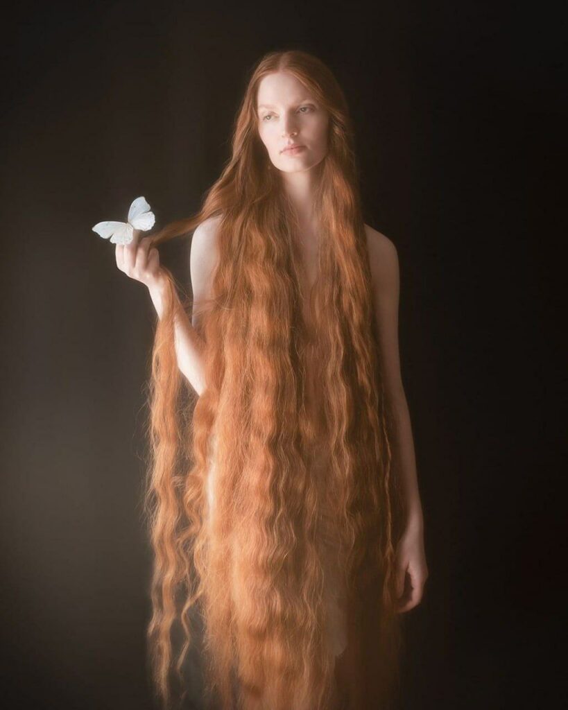 une femme rousse dont les cheveux descendent plus bas que ses genoux, un papillon blanc posé sur sa main