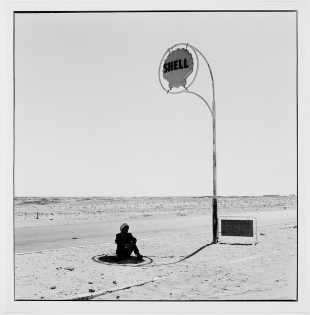 un algérien assis dans un paysage désertique à l’ombre de l’enseigne de la compagnie pétrolière, à l'ombre des stations service en fleurs
