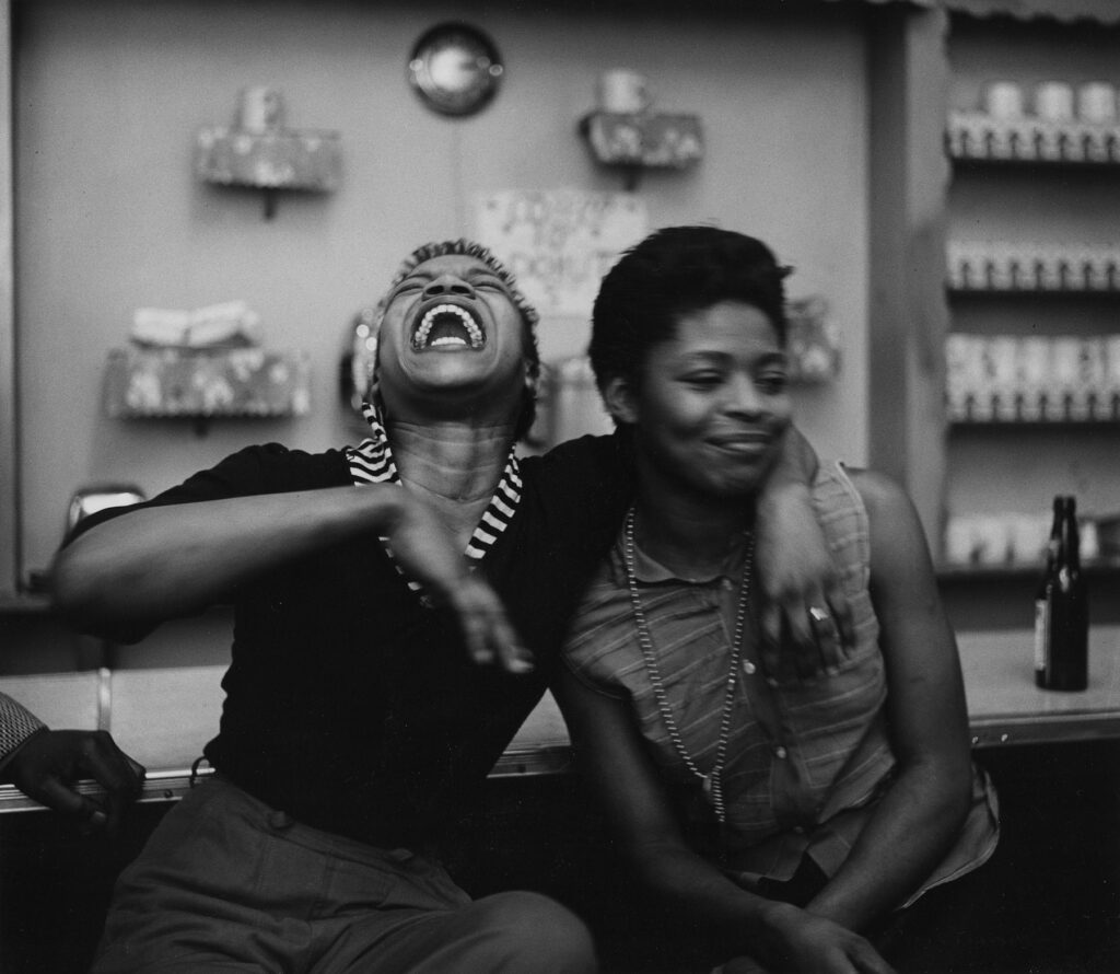 une femme noire, le bras passé autour du cou d'une amie, rit à gorge déployée, dévoilant sa superbe dentition