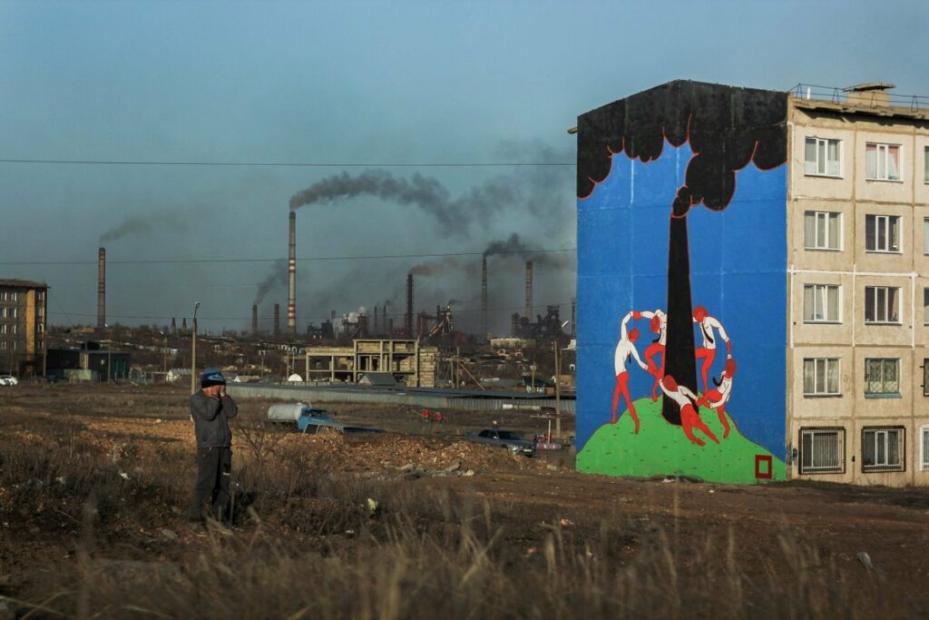 un ouvrier debout sur la friche devant une fresque murale représentant des travailleurs faisant une ronde joyeuse autour d'une cheminée d'usine, les vraies cheminées crachant une fumée noire à l'arrière plan