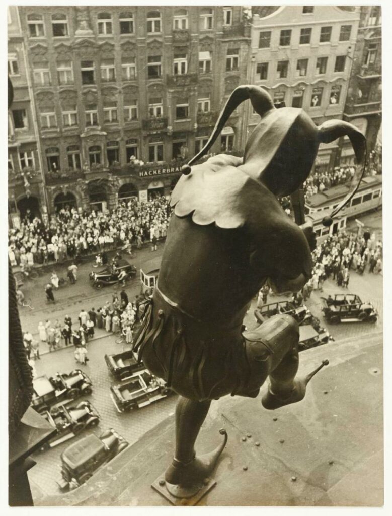 vue prise en 1934 depuis la tour de l'Hôtel de Ville de Munich surplombant la Marienplatz. Tritschler, positionné avec son appareil photo derrière le carillon historique de la tour,  regarde directement la foule dans la rue par-dessus l'épaule du fou