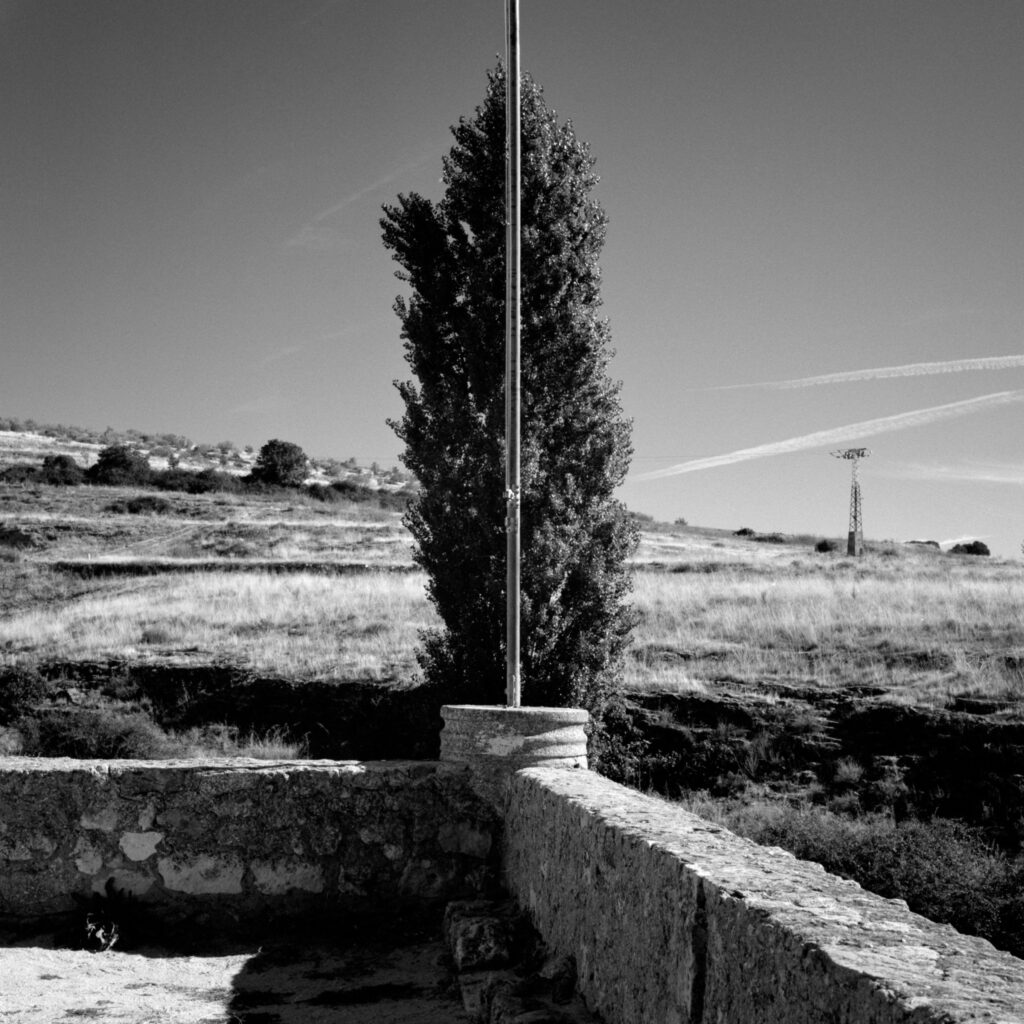 sur une terrasse donnant sur une colline déséchée, un poteau sépare en deux moitiés un cyprès (ou un if) au second plan