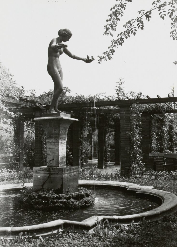 statue de femme aux bras ouverts dans un jardin, des oiseaux posés sur ses mains