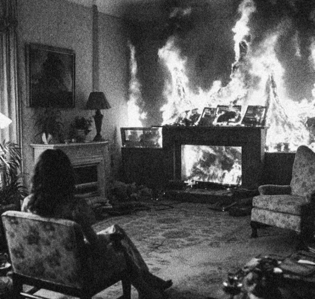 assise dans son fauteuil, une femme regarde le feu dans la cheminée. les flammes sont en train de gagner le reste de la pièce