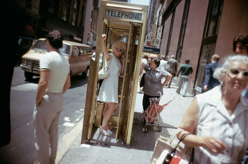 une femme, le bras levé dans une cabine téléphonique, dans le centre ville en plein soleil