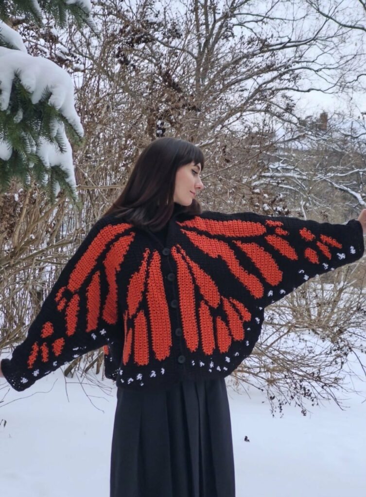 les pieds dans la neige, une jeune femme présente son cardigan papillon fait main