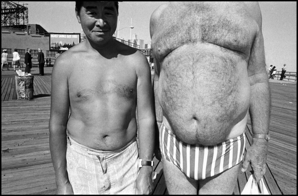 Deux hommes, dont l'un en surpoids, de grande taille et à la tête hors cadre, exhibent leur ventre en prenant un bain de soleil sur la promenade